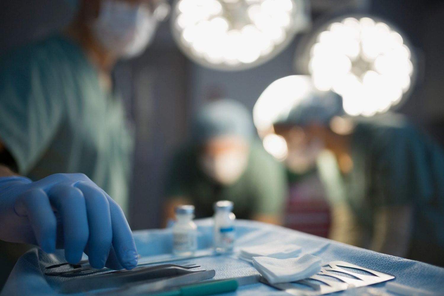 Surgeon reaching for scalpel during surgery. Drugs in foreground, medical assistants in background.