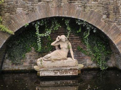 Stone statue of a reclining woman under an ivy-covered arch by water.