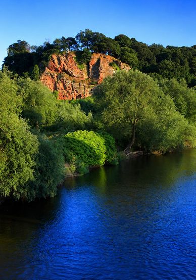 Sunlit red cliffs and lush greenery by a calm blue river under a clear sky.