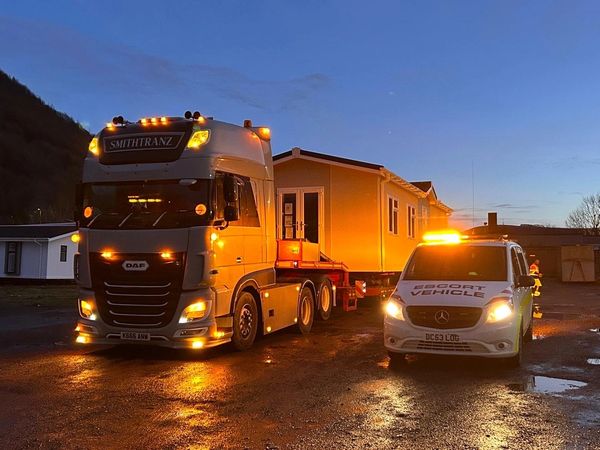 A large truck and recovery vehicle transport a modular home at dusk.