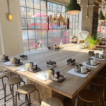 Sunlit rustic workshop table set with stools and small bottles near a large window.
