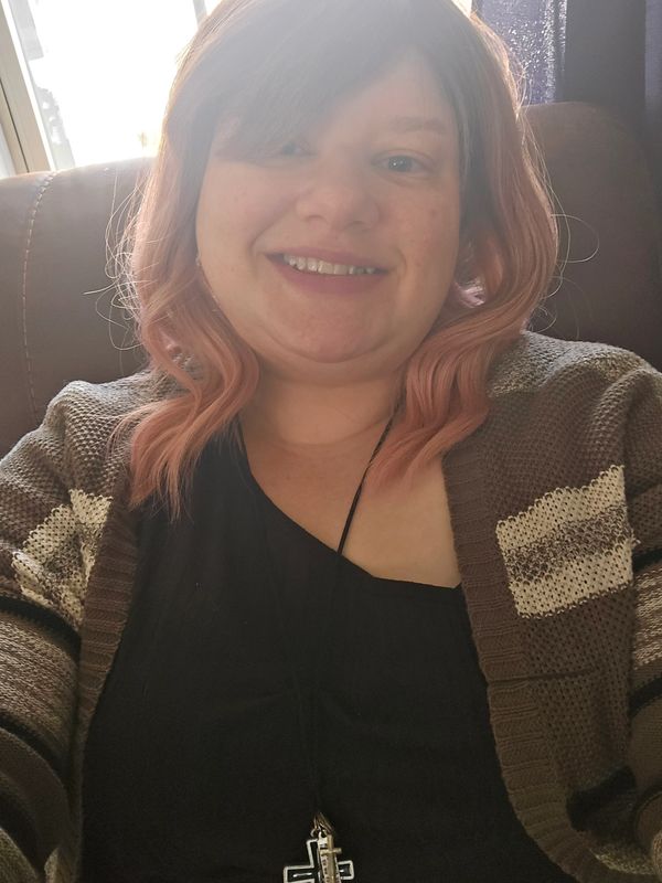 Smiling woman with pink hair wearing a striped cardigan and cross necklace.