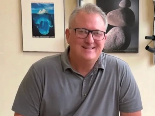 A smiling man wearing glasses and a grey polo shirt indoors.