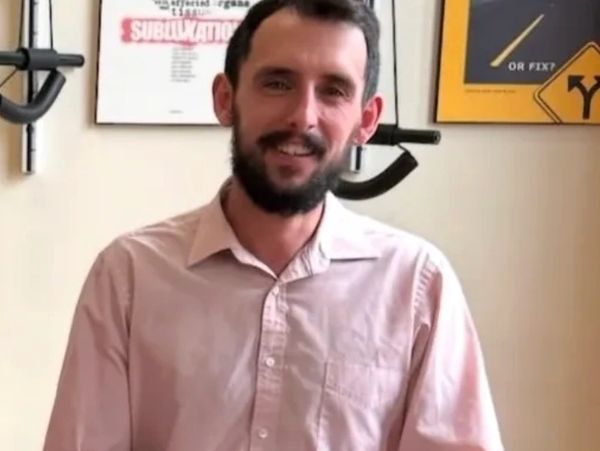 Man with beard wearing pink shirt smiling indoors.