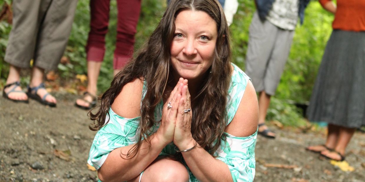 Woman with long hair kneeling and smiling with hands in prayer pose outdoors.