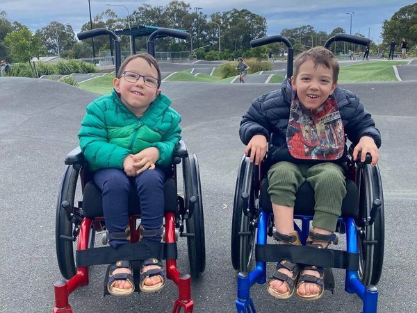 Two smiling boys in wheelchairs enjoy time outdoors at a park, wearing jackets and sandals.