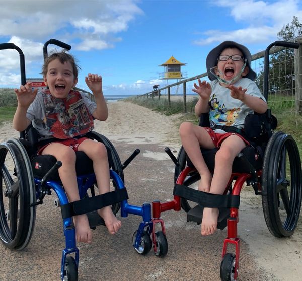 Two joyful boys in wheelchairs hold hands and smile while on a path leading to the beach.