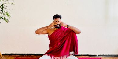A man in a red shawl practices yoga covering his eyes with his hands.