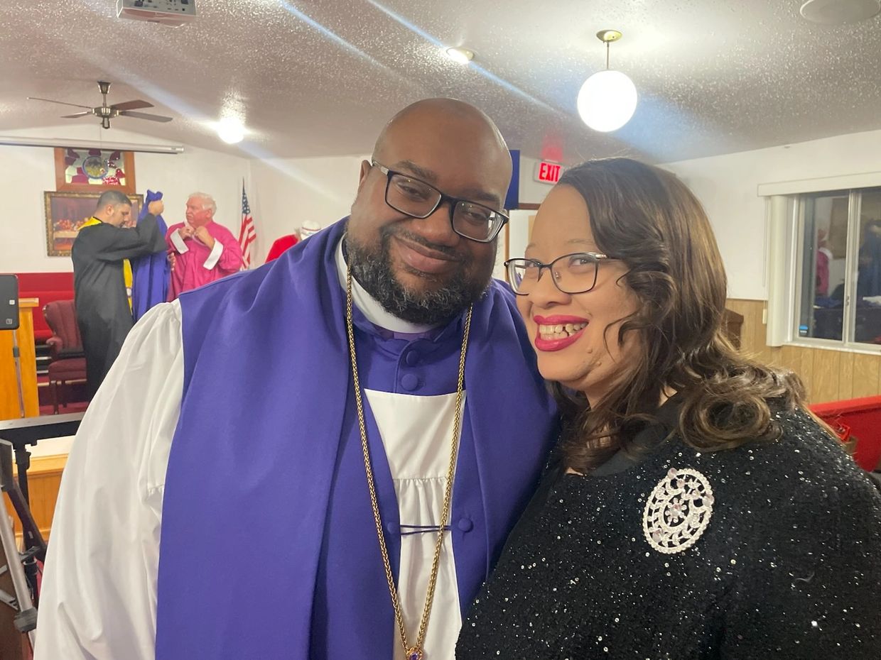 A smiling man in religious attire and a woman with a brooch pose together indoors.