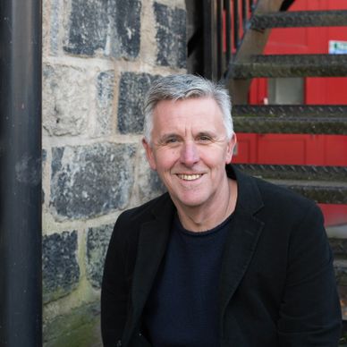 Smiling man in black jacket sitting outdoors by stone wall and stairs.