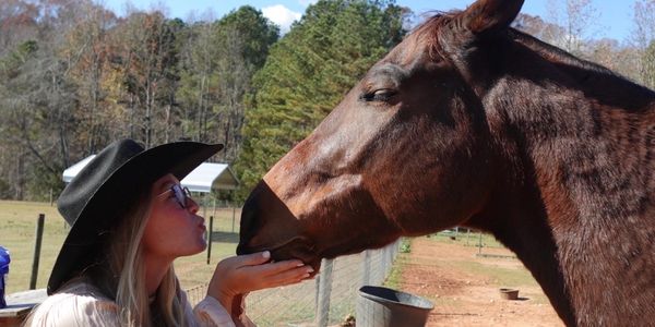 A woman in a black hat kisses a horse on the nose outdoors.