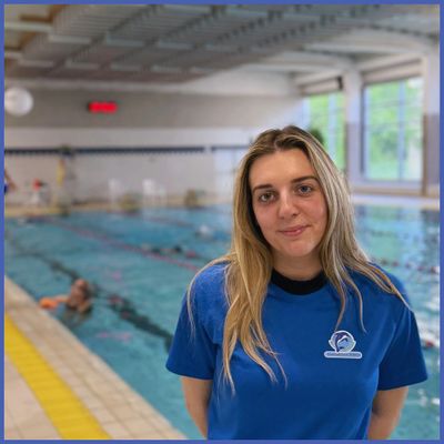A woman in a blue shirt stands by an indoor swimming pool.