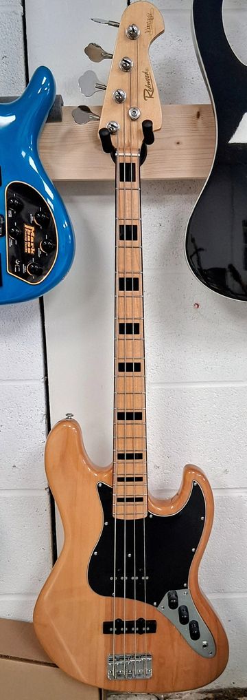 A natural wood finish electric bass guitar with black pickguard hanging on a wall.