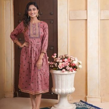 A woman in a pink traditional dress poses indoors beside a flower vase.
