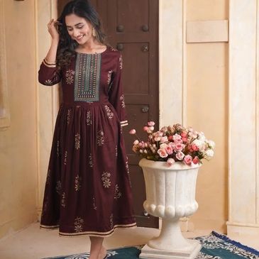 Woman in a maroon embroidered dress smiling indoors with a floral vase.