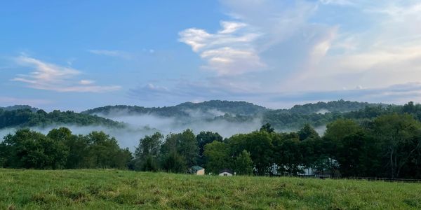 A bright blue sky, vibrant green mountains, and fog and clouds can be seen from this hilltop. 