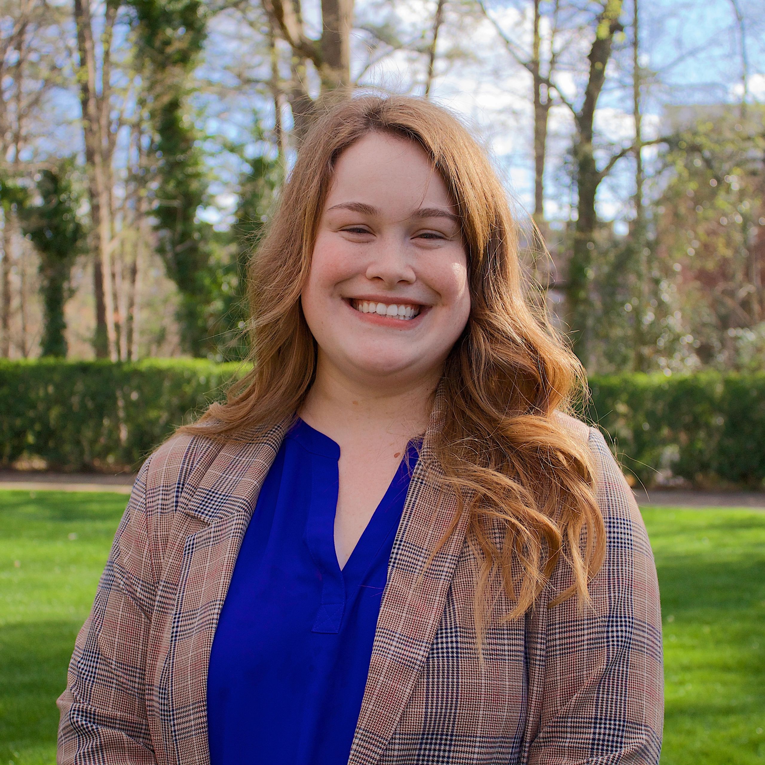 This picture is of Marissa Hall with greenery behind her in a blue shirt and plaid blazer.