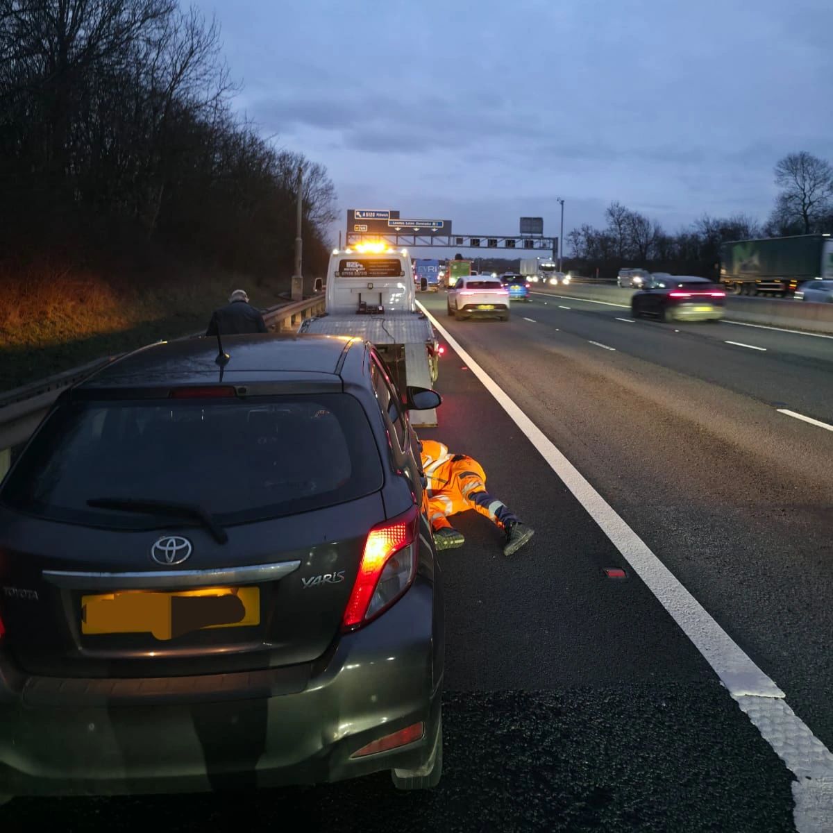 Tow truck operator working under a Toyota Yaris on a busy highway at dusk.