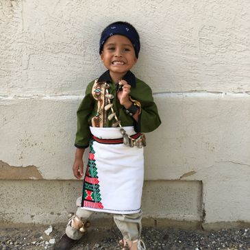 A young boy smiling in traditional attire with a decorated apron and headband.