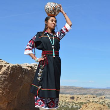 Woman in traditional attire carries a decorated pot on her head against a clear sky.