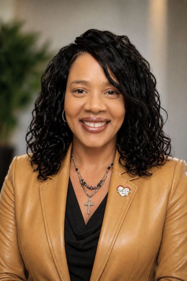 Smiling woman with curly hair wearing a tan blazer and cross necklace.