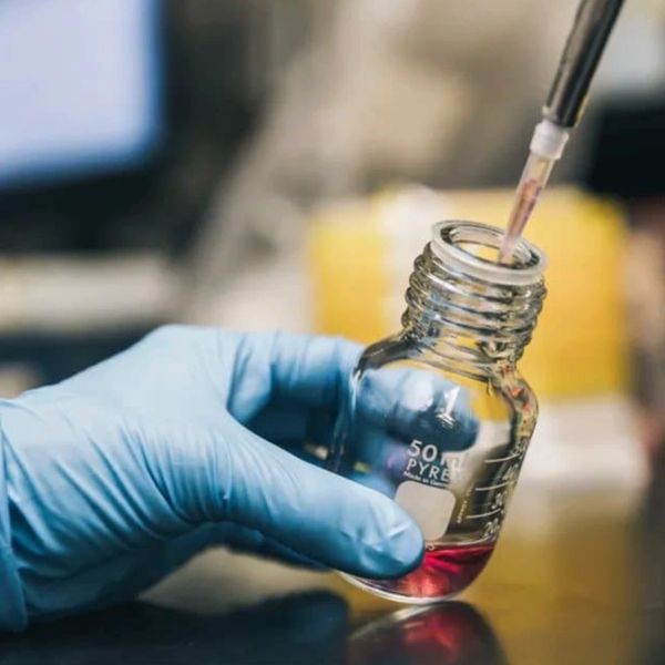 Gloved hand holding a glass bottle with a dropper in a lab.