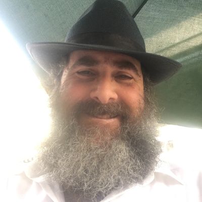 Smiling man with a thick beard and black hat under shaded light.