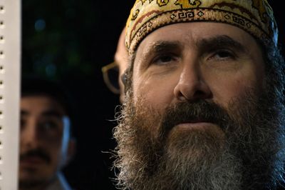 Close-up of a bearded man wearing a patterned headscarf with two other men blurred in the background.