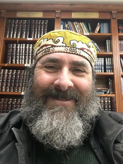 A man with a gray beard wearing a colorful cap smiles in front of a bookshelf.