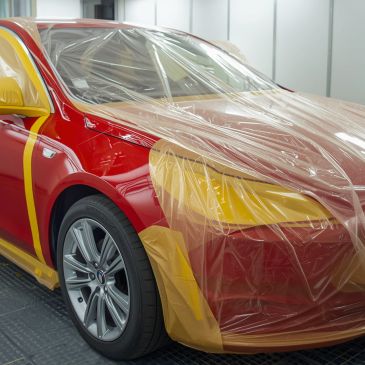 Red car partially covered with protective plastic and masking tape in a workshop.