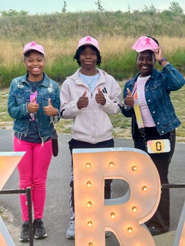 Three people giving thumbs up, wearing pink accessories for breast cancer awareness.