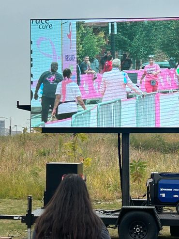 Large screen showing people at a breast cancer awareness event outdoors.