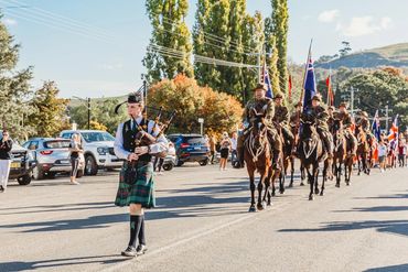 Man in kilt playing bagpipes leading a parade of horse riders with flags.