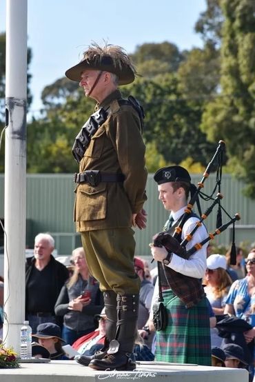 A man in historical military attire stands solemnly next to a bagpiper in traditional Scottish dress.