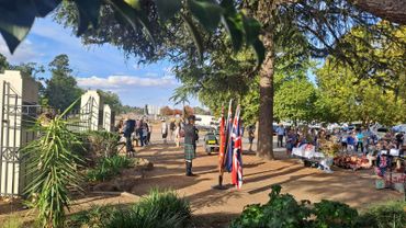 A man in a kilt plays bagpipes at an outdoor event with flags and a crowd gathered.