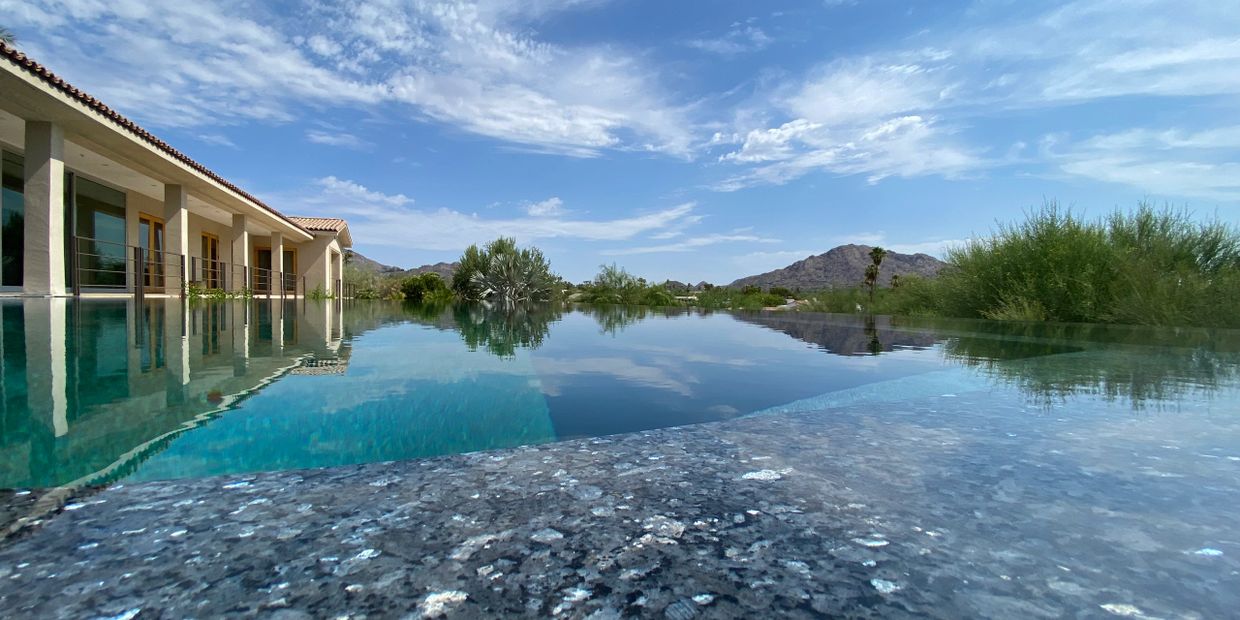 Infinity pool reflecting sky and surrounding landscape beside a modern house.