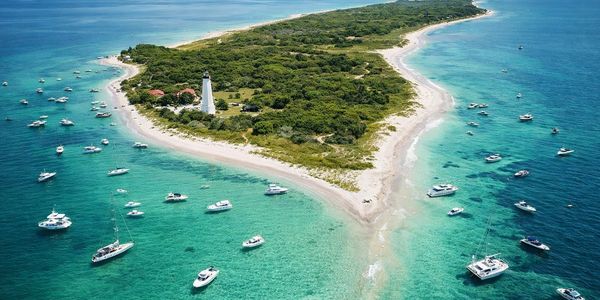 A lush island with a lighthouse surrounded by turquoise waters and numerous boats.