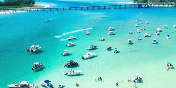 Boats anchored in clear turquoise water near a sandy beach with people enjoying the shallow sea.