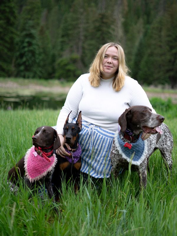 A woman in a white sweater poses with three dogs wearing colorful bandanas in a grassy field.