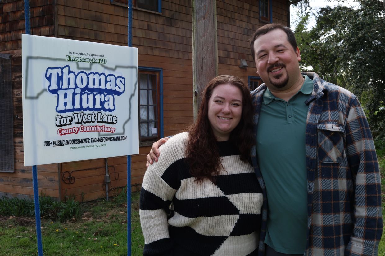 Two people smiling beside a campaign sign for Thomas Hiura.