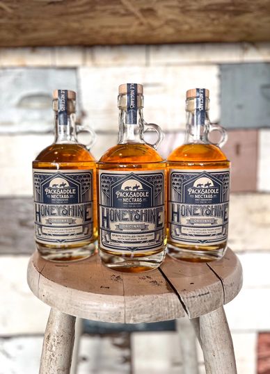 Three bottles of Pack Saddle Nectars Honeyshine on a wooden stool.