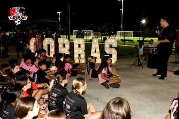 Youth soccer team listening to a coach at night with illuminated COBRAS sign.
