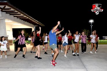 A group of young soccer players and a coach dancing outdoors at night.
