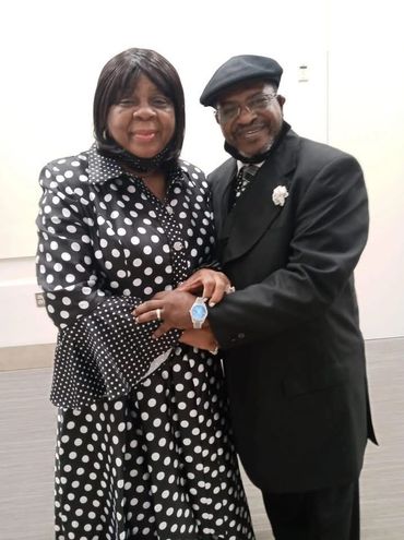 A smiling couple dressed in black and white, with the woman in a polka dot dress and the man in a suit and cap.