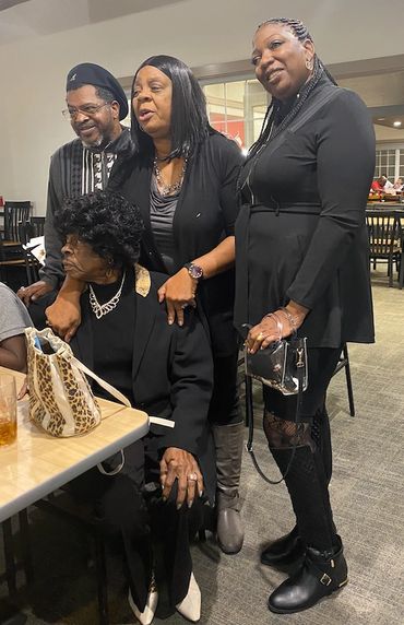 Four people posing together indoors, three standing and one seated, dressed in black.