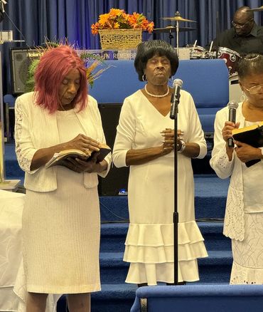 Three women in white dresses praying and reading from books in a church.