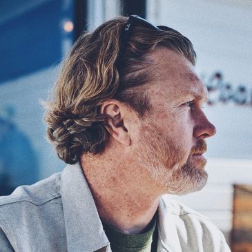 Man in light jacket holding a glass of beer at an outdoor wooden table.