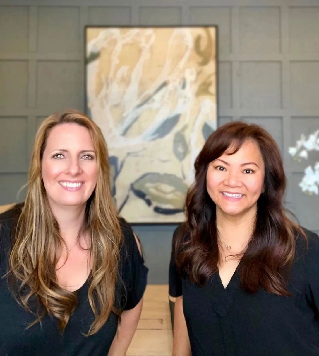 Two smiling women with long hair pose indoors in front of abstract wall art.