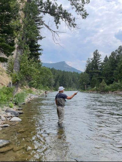 Person fishing in a river surrounded by trees and mountains.