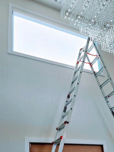 Tall ladder leaning against a wall near a large window and chandelier.